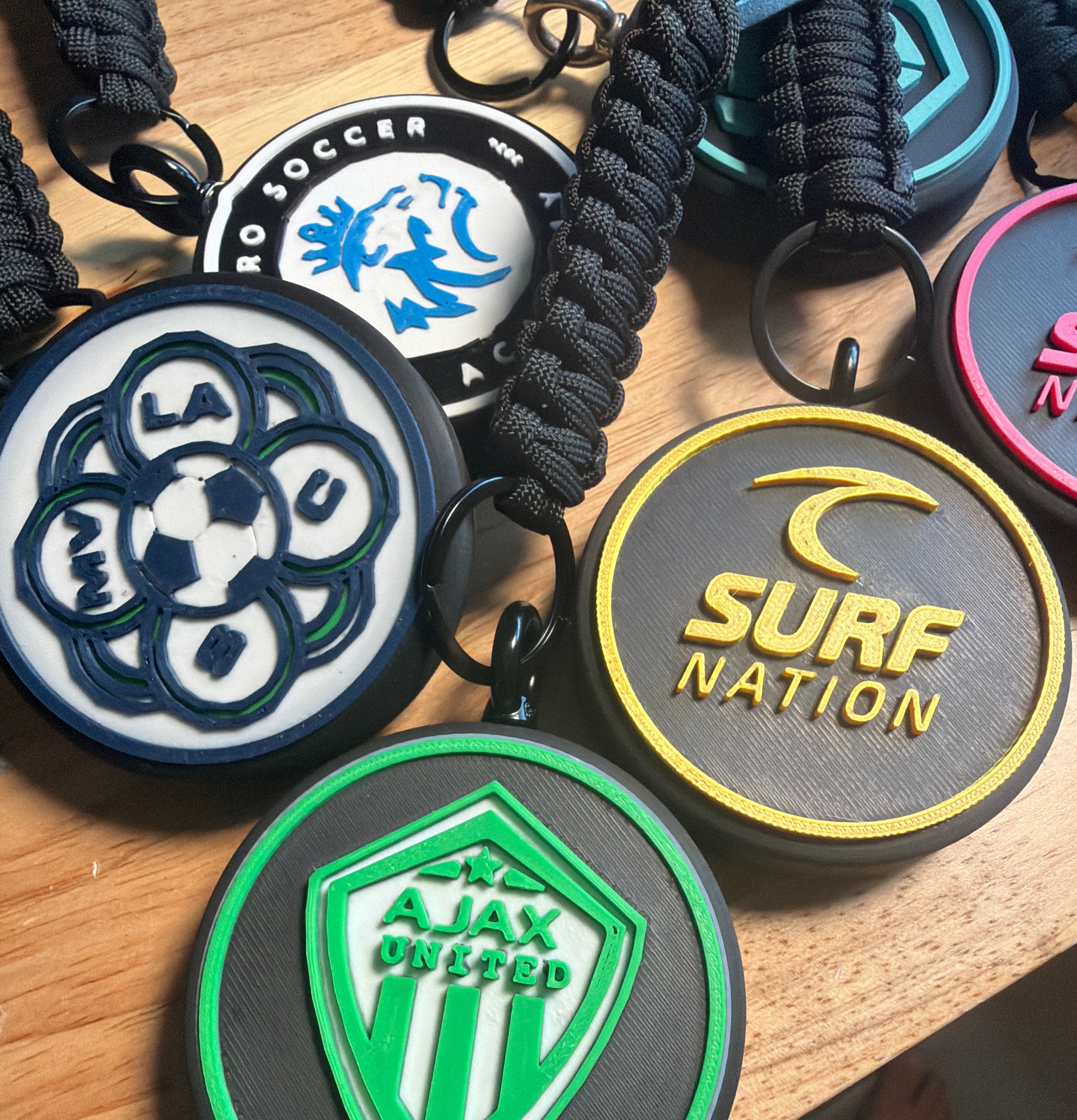 Custom Tracker Tag examples for Ajax United, Surf Nation, and SC Pro Soccer with black braided paracord attachments