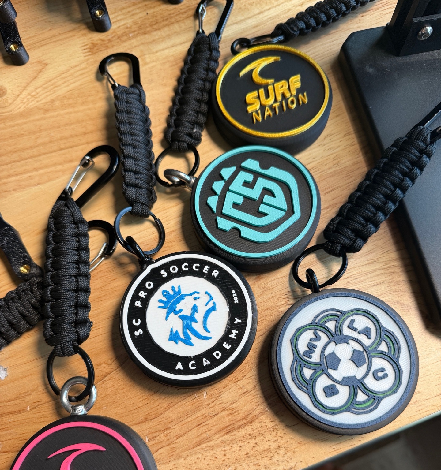 Real custom Tracker Tag examples with Surf Nation, CS Futsal, and SC Pro Soccer Academy team logos beside black braided paracord attachments
