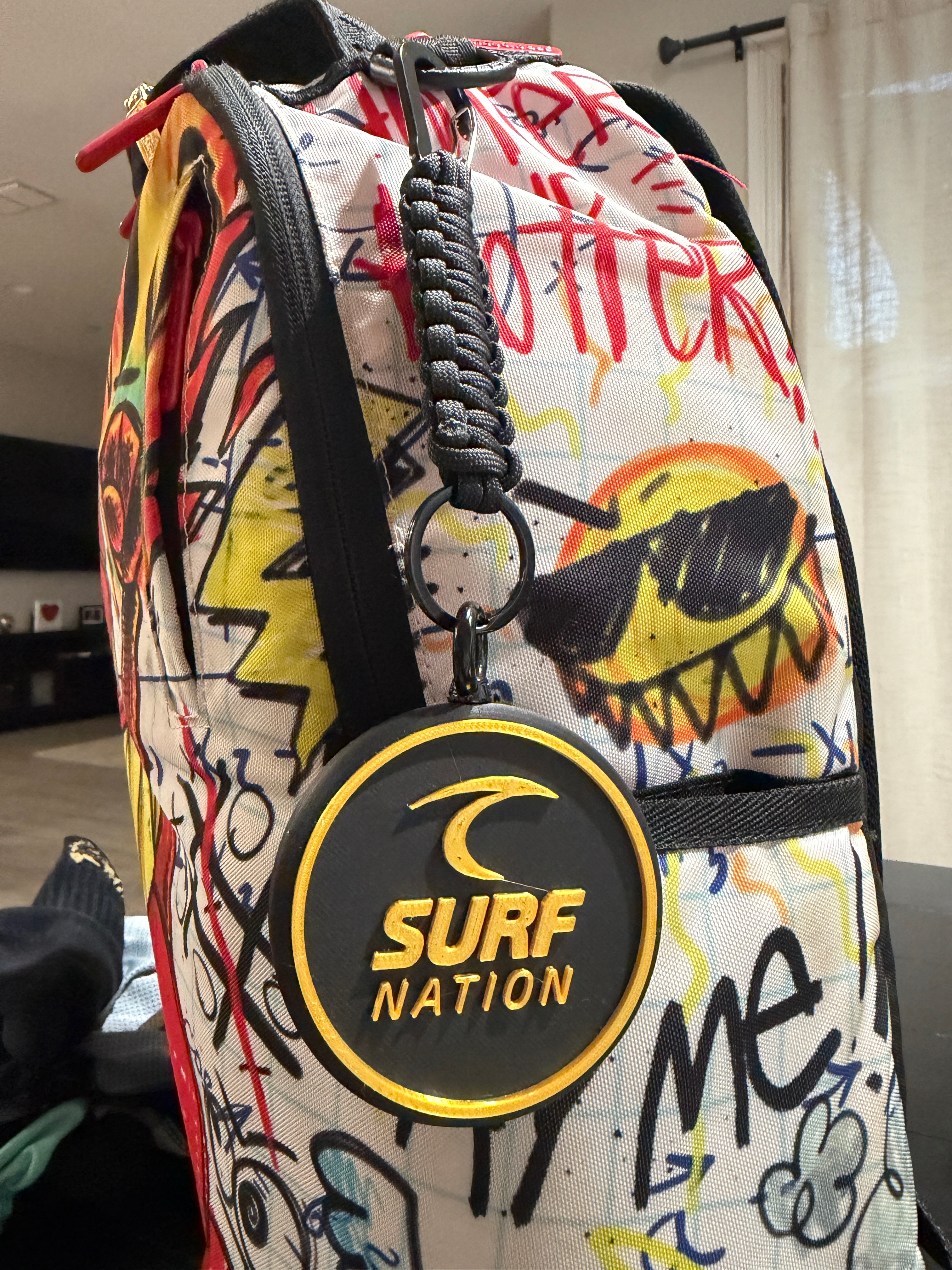 Gold Surf Nation Tracker Tag attached to a colorful backpack with a detailed all-black braided paracord, black carabiner, and split ring